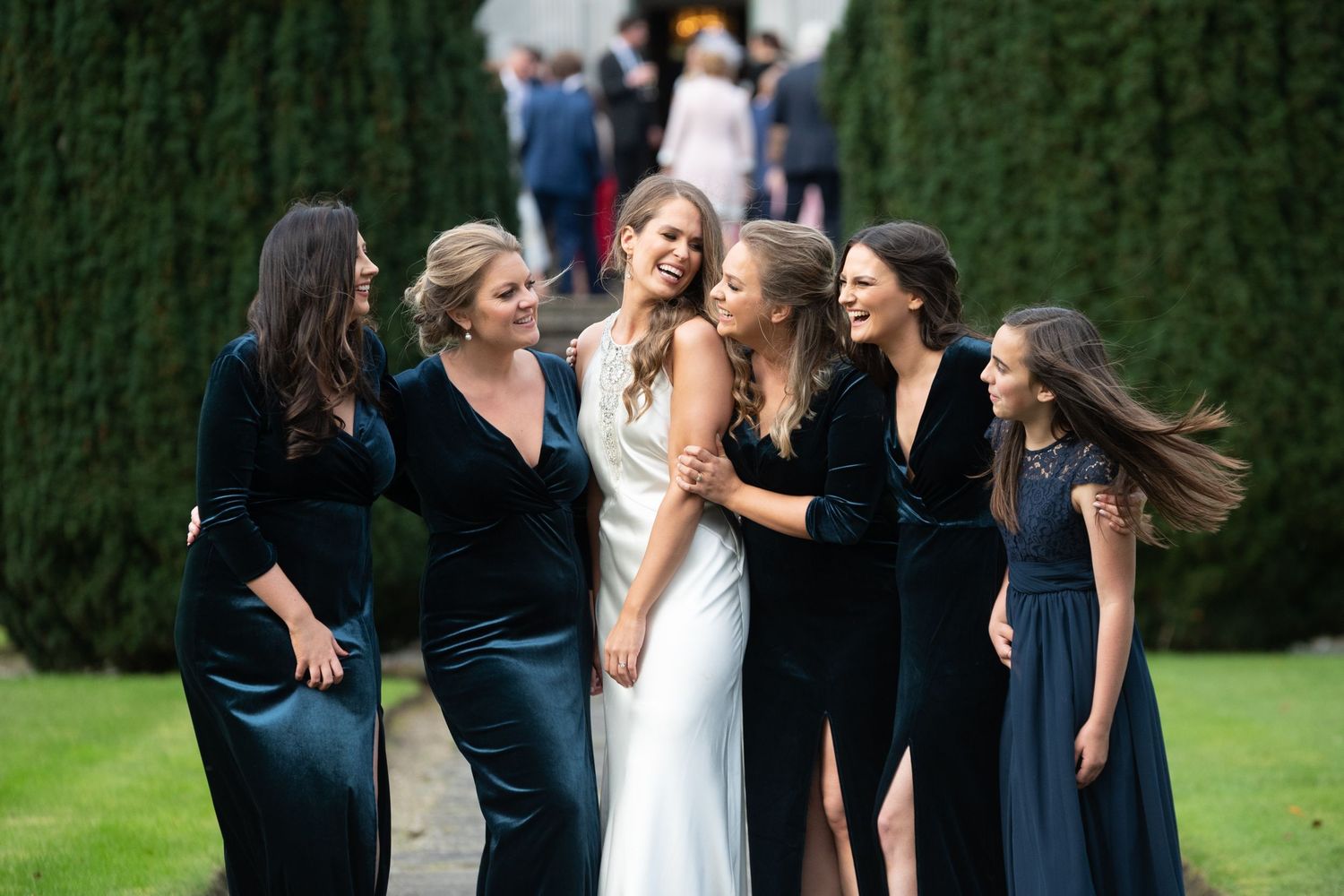 Bride in white gown laughing with bridesmaids in dark velvet dresses outdoors at elegant wedding venue.