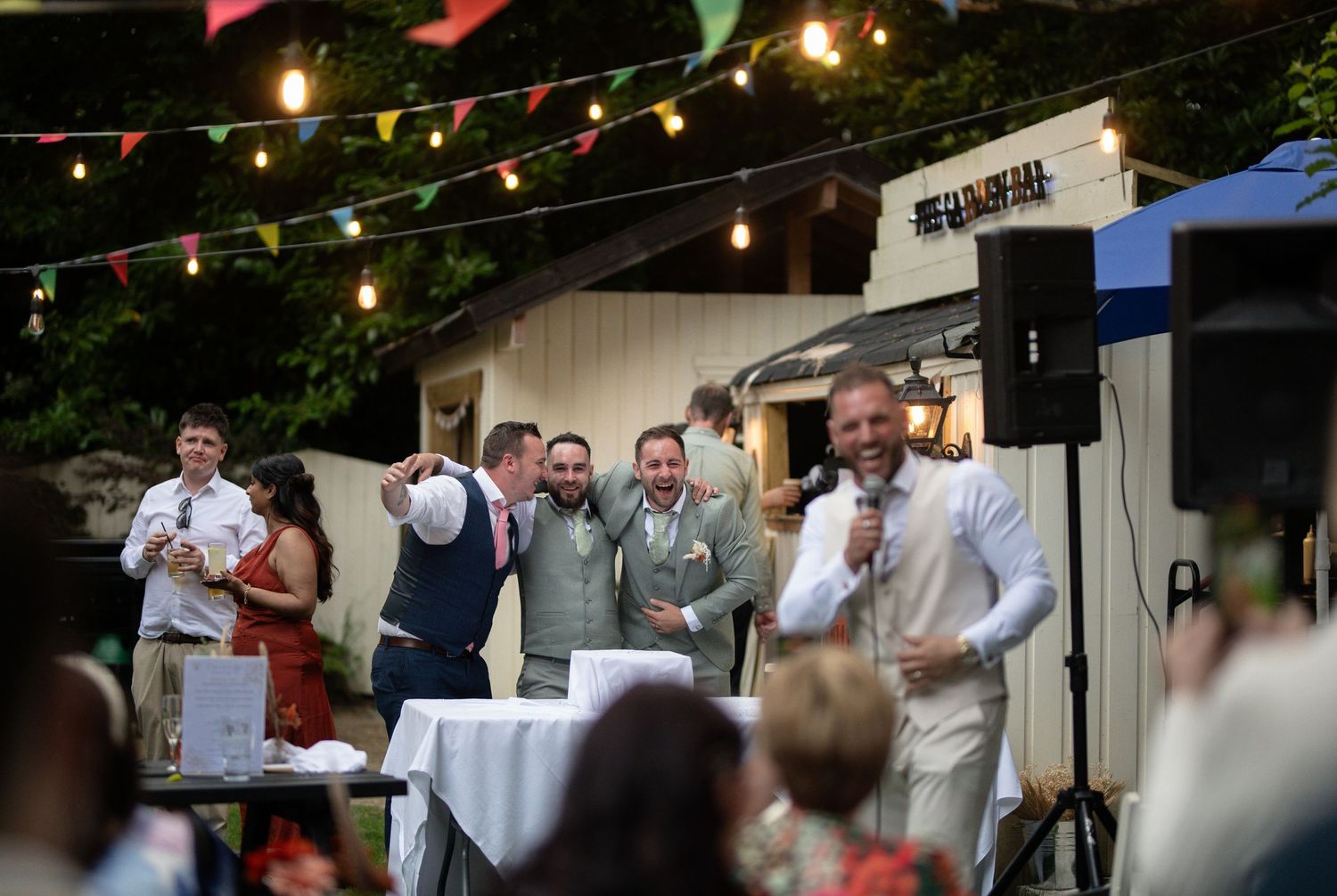 Cheerful wedding guests and groomsmen laughing during speeches at a festively lit outdoor reception.
