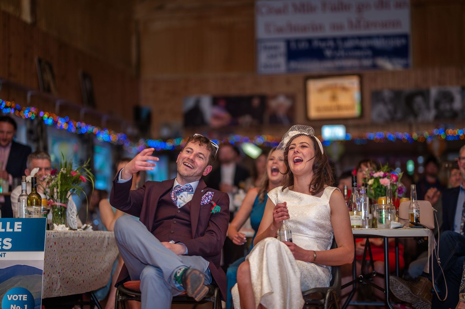 Bride and groom laughing joyfully at wedding reception, seated at decorated table with colorful bunting in rustic venue.