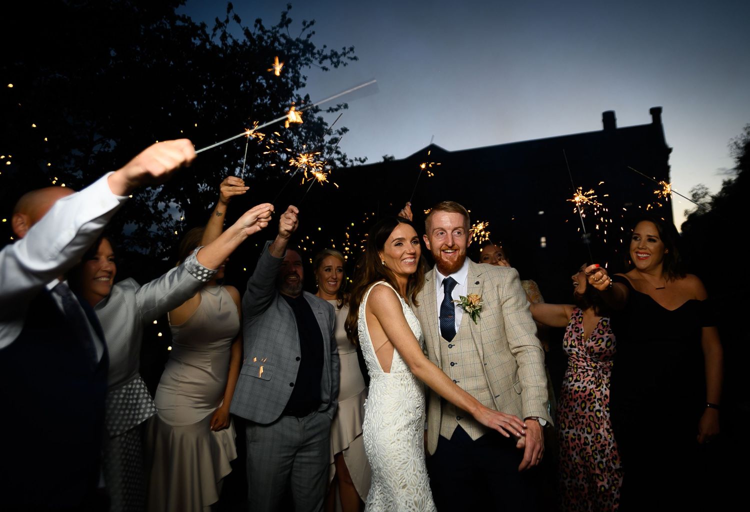 Newlywed couple walking through sparkler tunnel surrounded by wedding guests at twilight celebration.