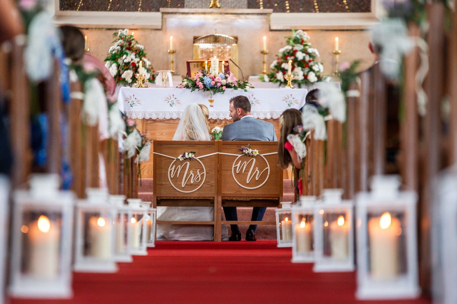Bride and groom seated at church altar during wedding ceremony, flanked by candles and floral arrangements.