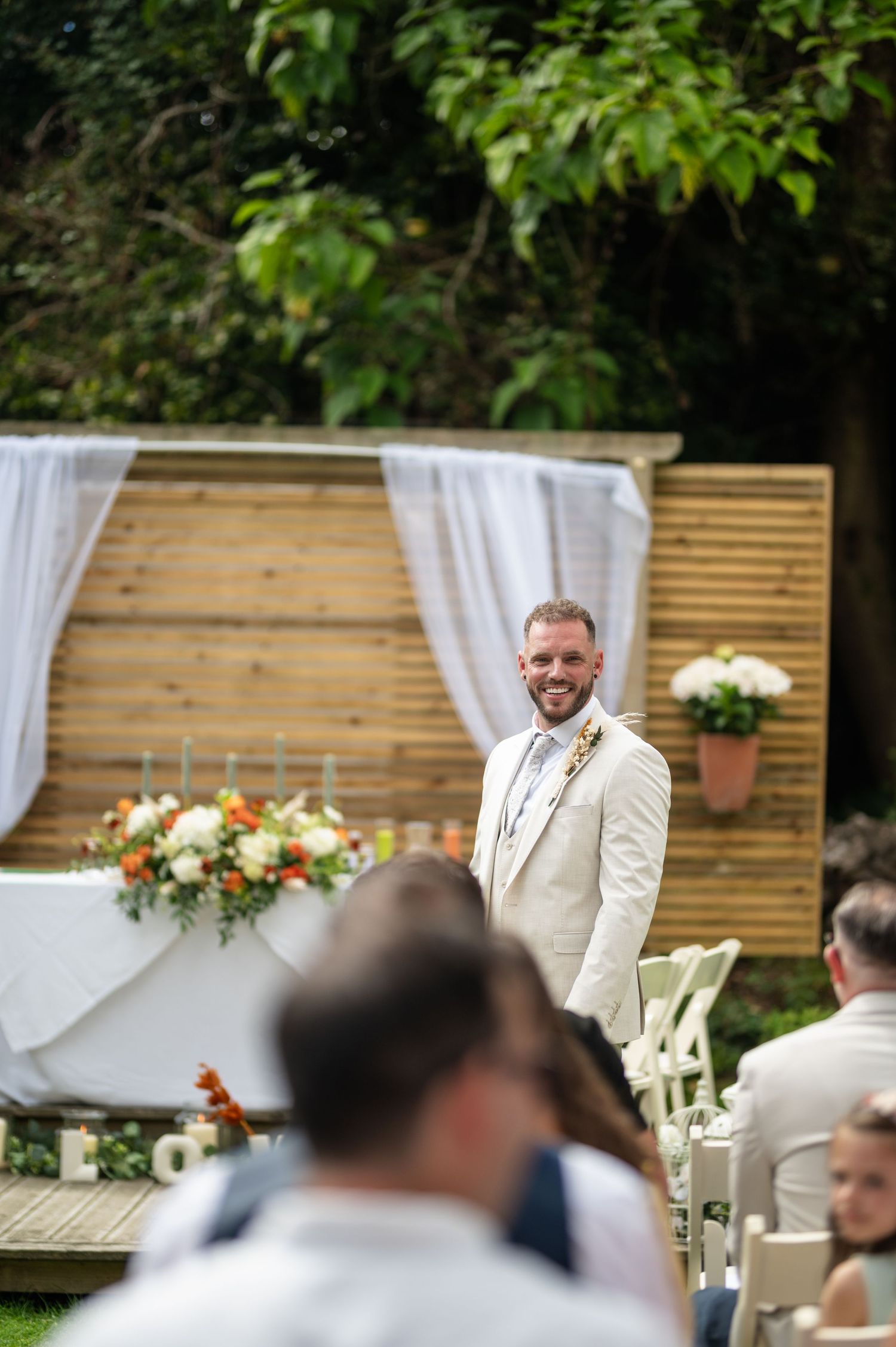 Smiling groom in cream suit standing at outdoor wedding ceremony altar.