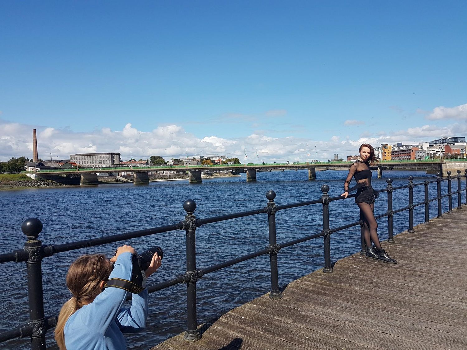 Photographer capturing a woman posing on a riverside dock with a bridge and blue sky backdrop.