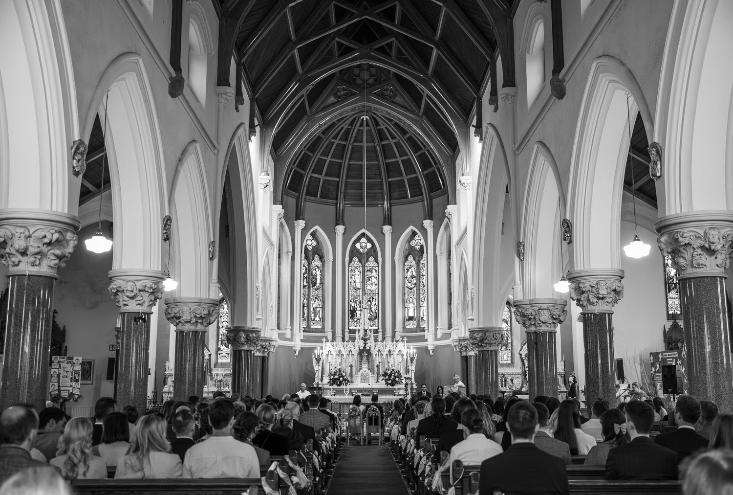 Black and white photo of wedding guests seated inside a grand church with arched ceilings and stained glass windows.