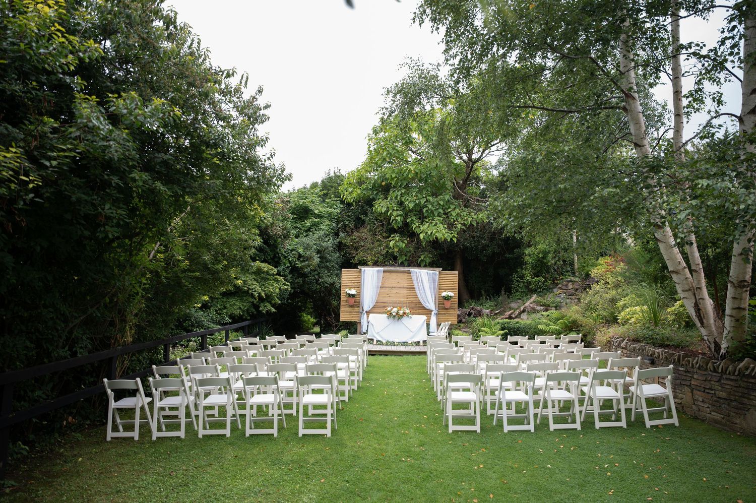 Outdoor wedding ceremony setup with white chairs on green lawn and floral arch backdrop surrounded by lush trees.