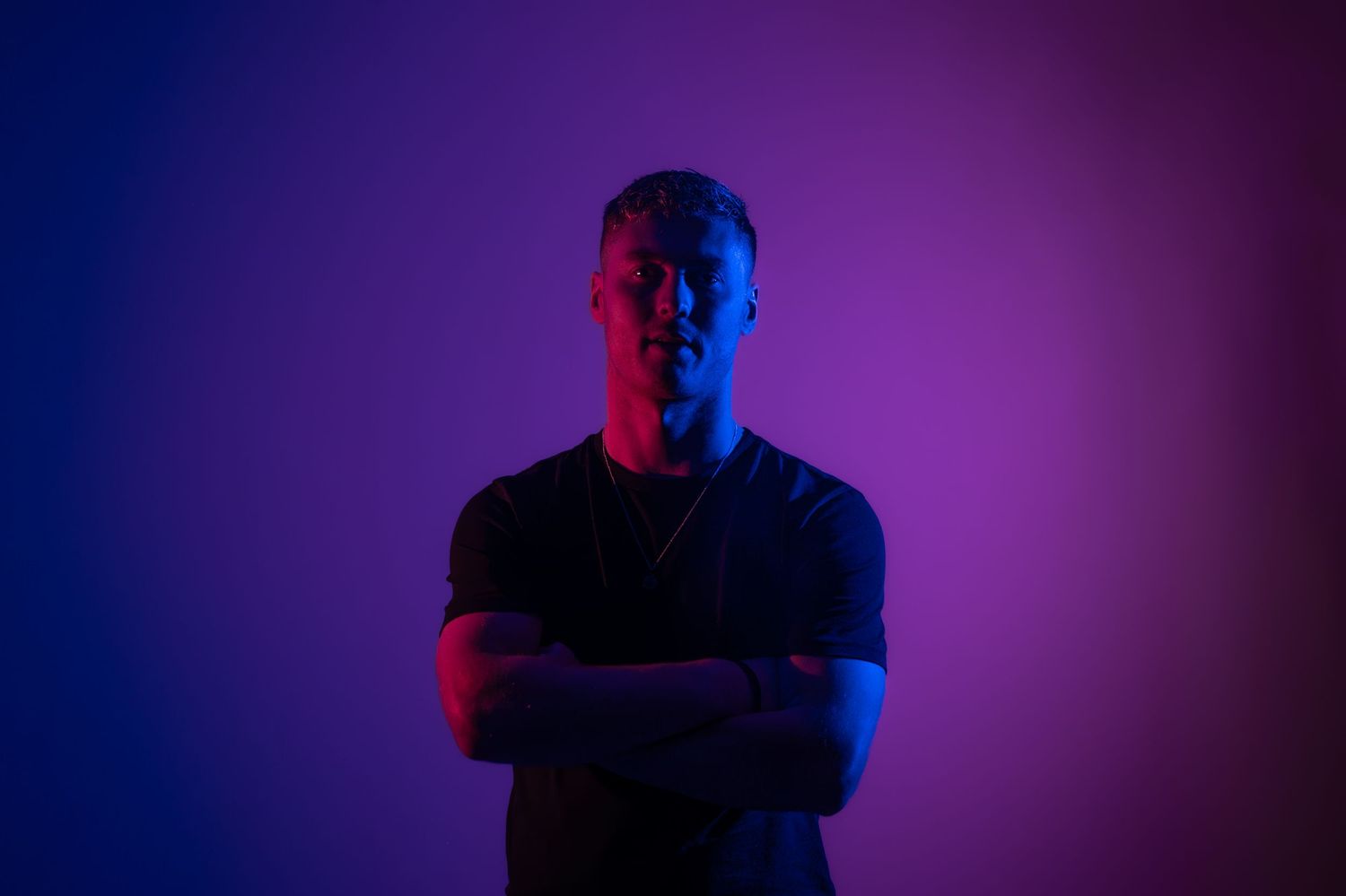 Man in black t-shirt with arms crossed under blue and red neon lighting.
