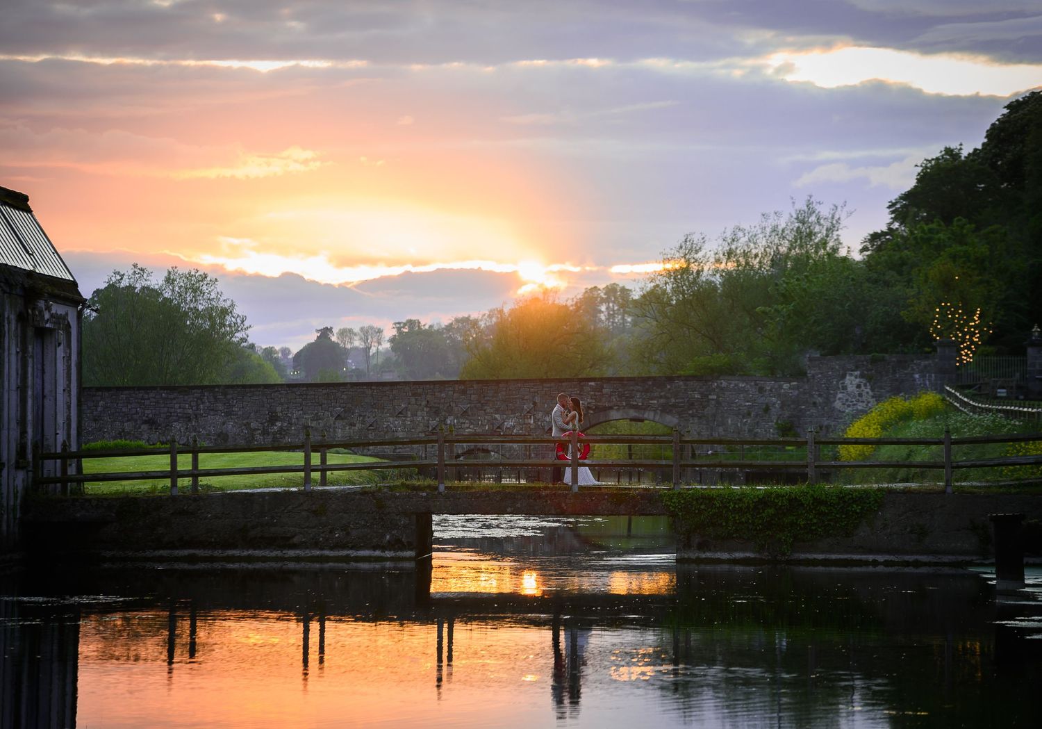 Couple on a stone bridge at sunset with golden reflections on calm water at a scenic Irish estate.