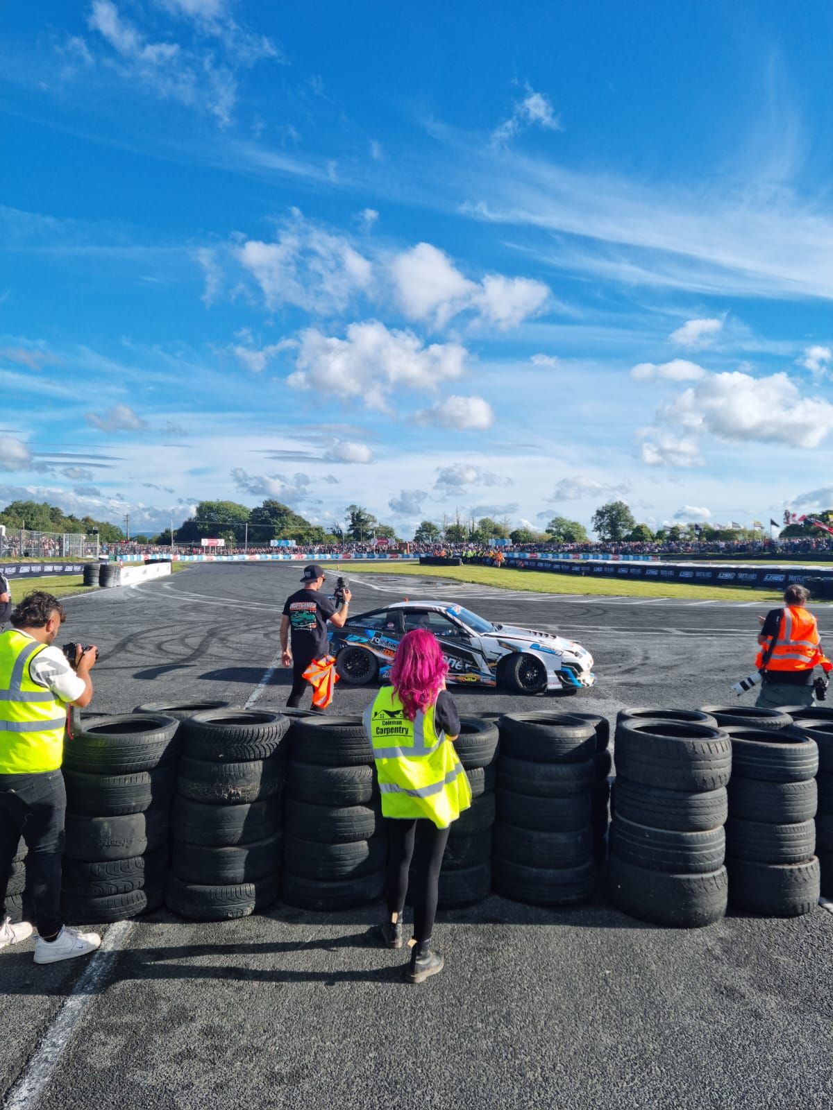 Pink-haired photographer shooting a race car drifting on a track surrounded by stacked tires.