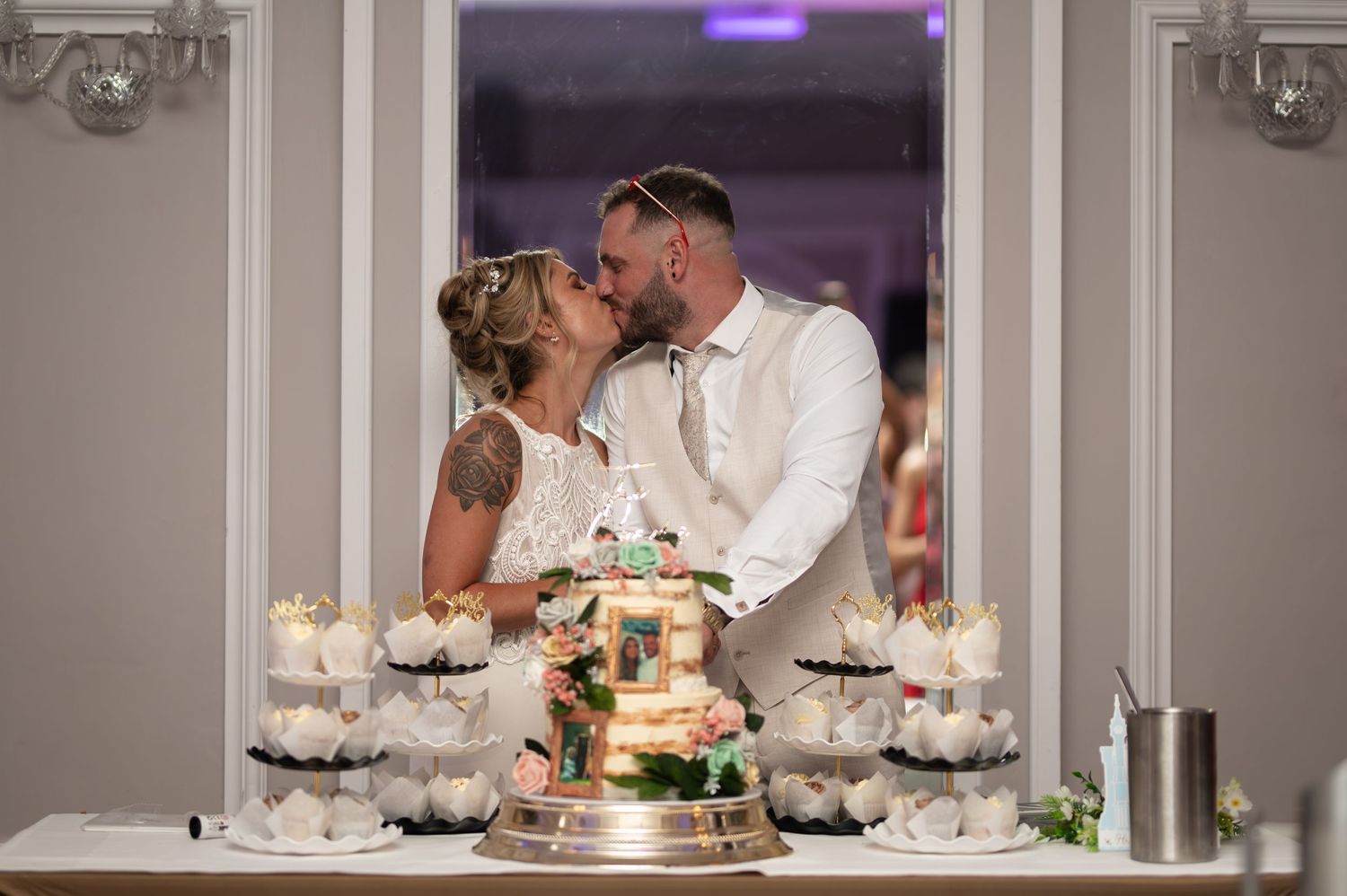 Bride and groom share a kiss behind a elegant wedding cake display with cupcakes at their reception.