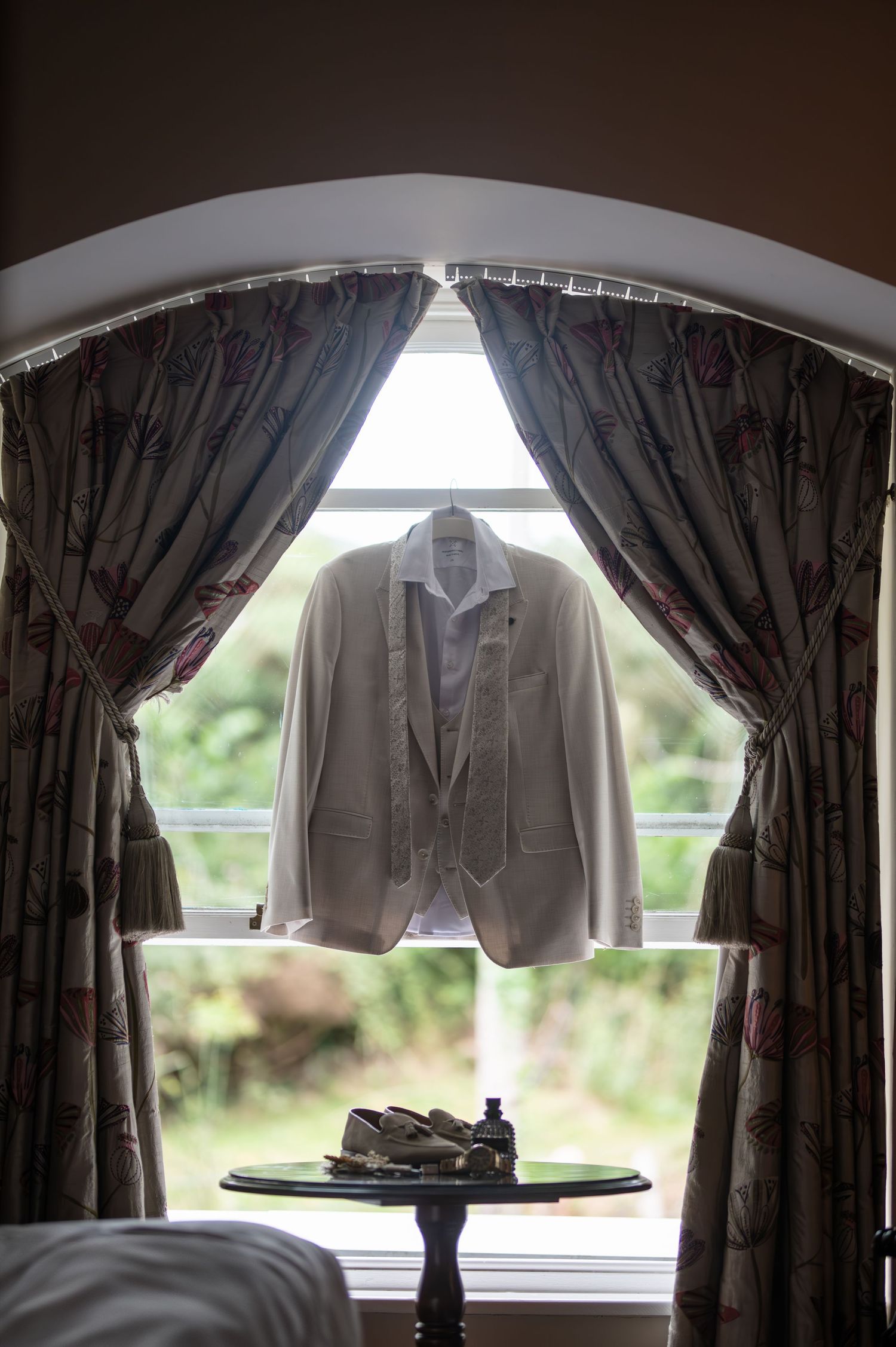 Groom's white suit jacket hanging in an arched window with floral curtains.
