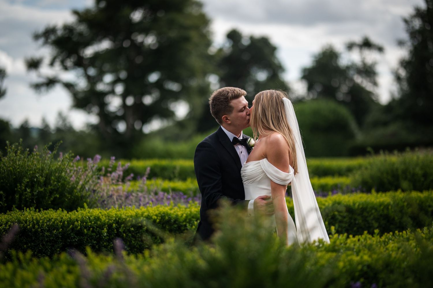 A bride and groom share a romantic kiss surrounded by lush green hedges and lavender in an elegant garden setting.