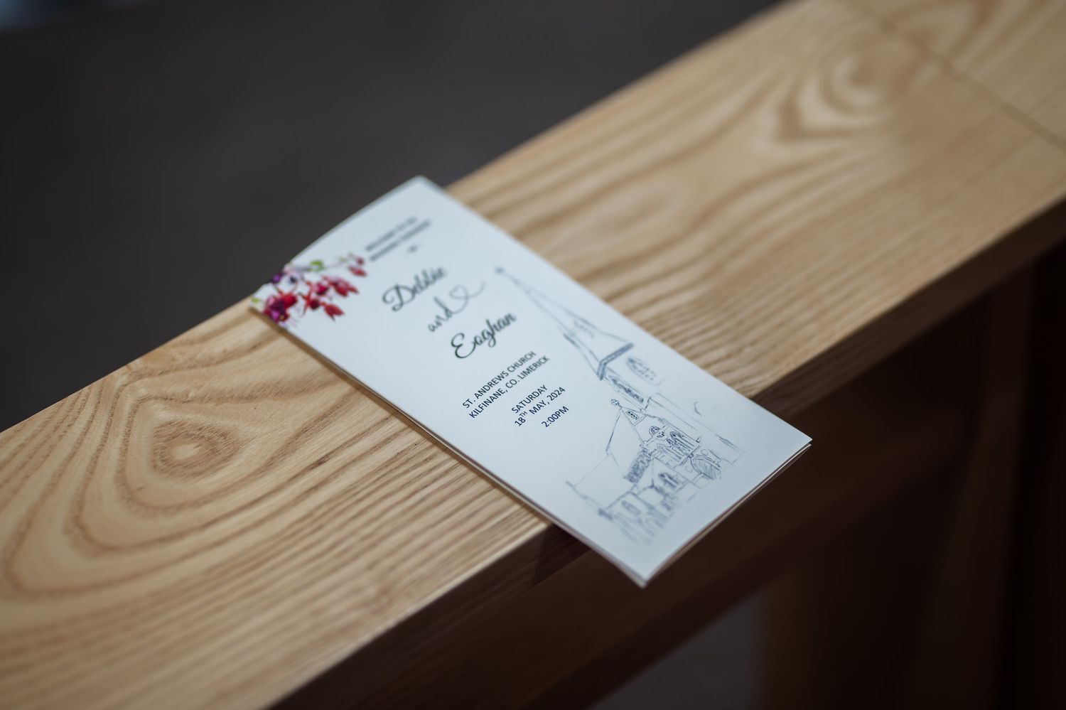 Wedding program card with red floral accents and church illustration resting on a wooden pew.