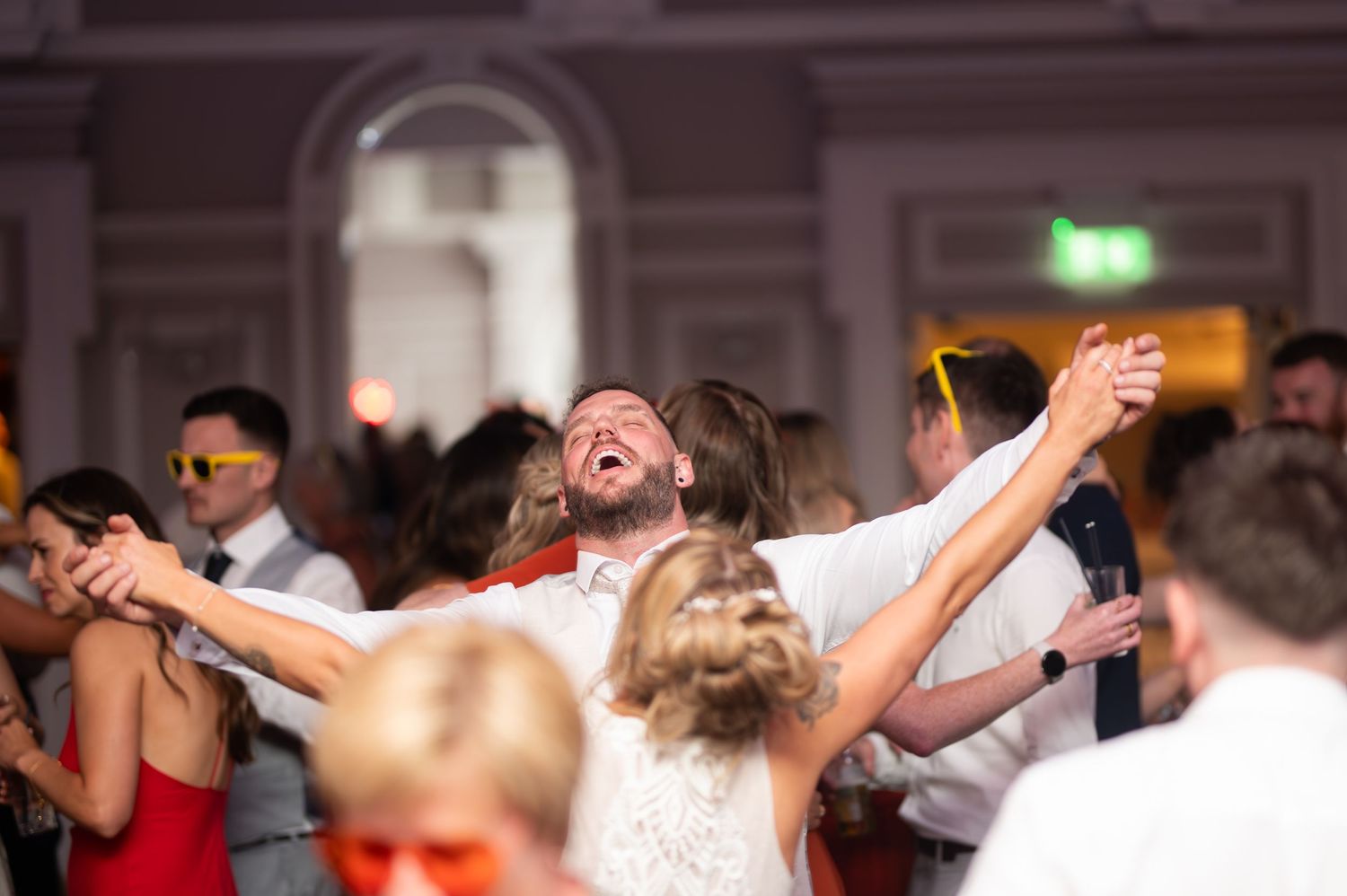 Joyful groom laughs with arms raised while dancing with guests at a lively wedding reception.