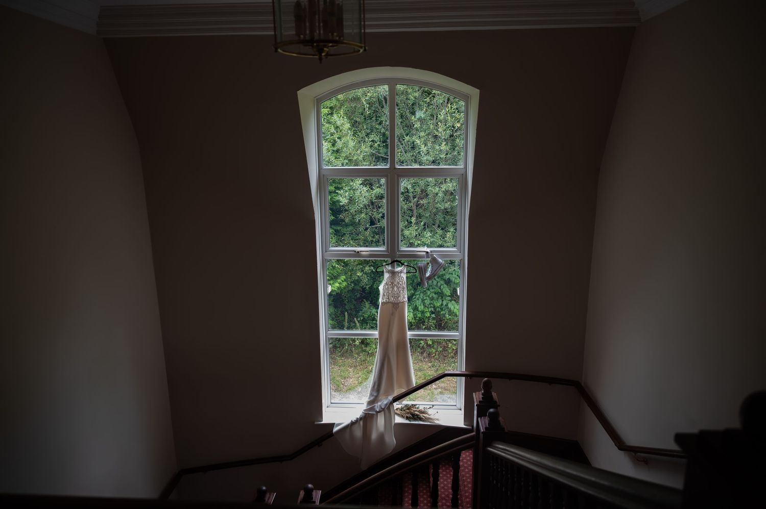 Elegant wedding dress hanging in arched window of a historic manor staircase.