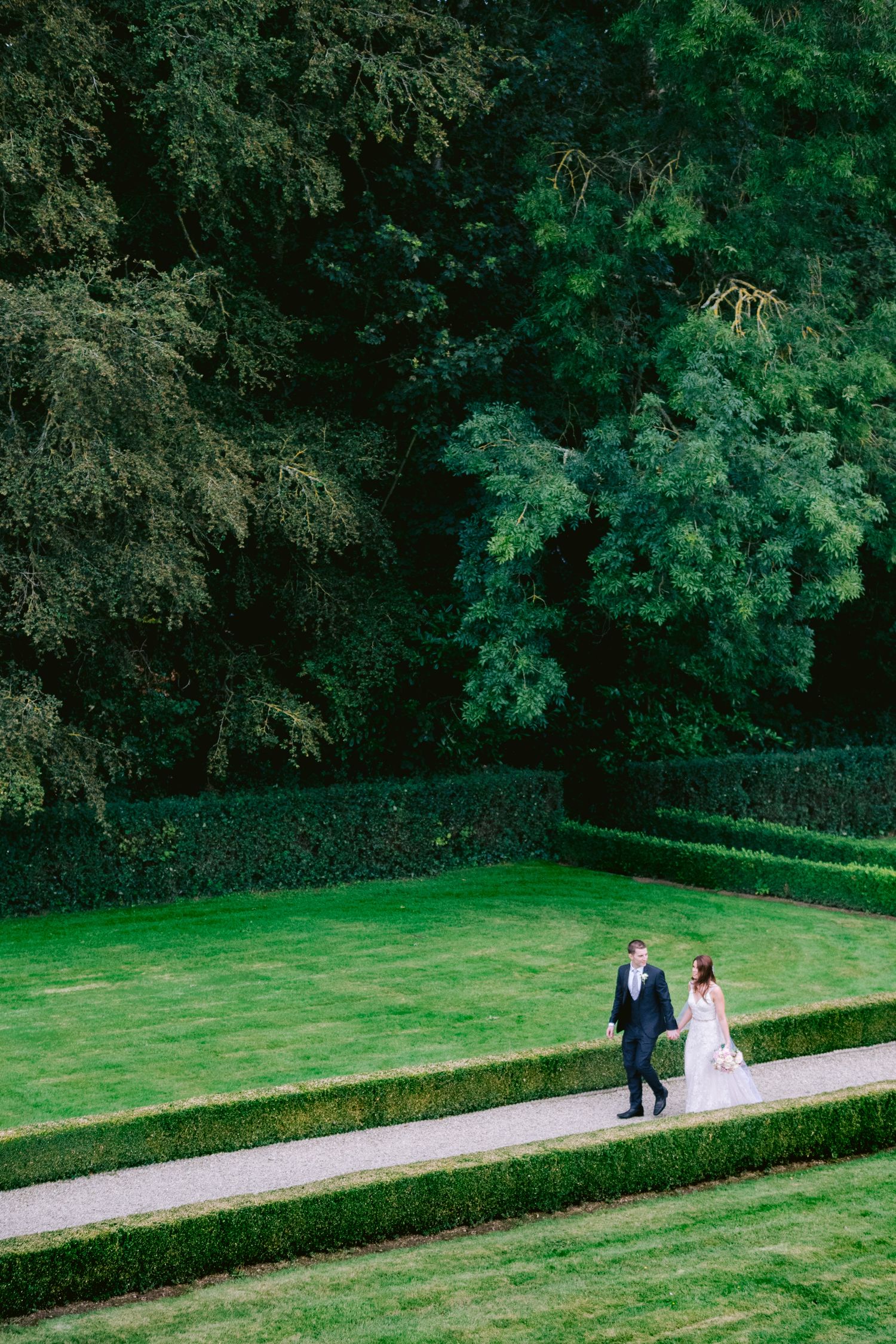 A bride and groom walk hand in hand along a garden path surrounded by manicured hedges and tall trees.