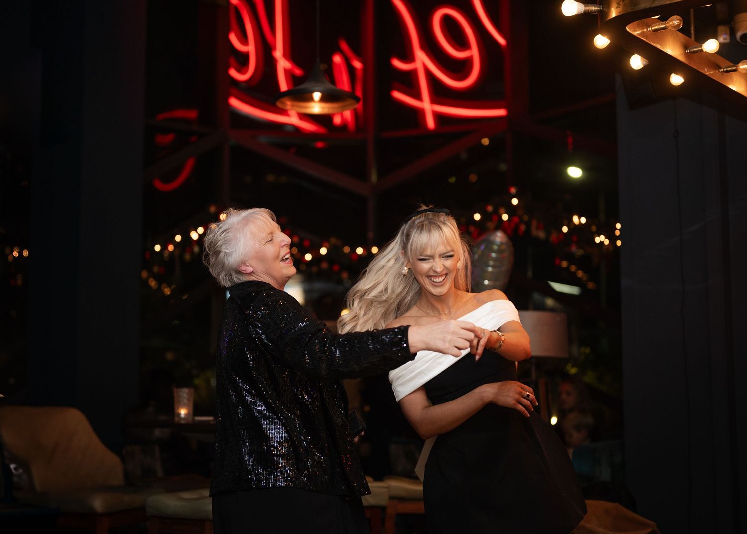 Two women laughing and dancing together at a stylish evening party with neon signs.