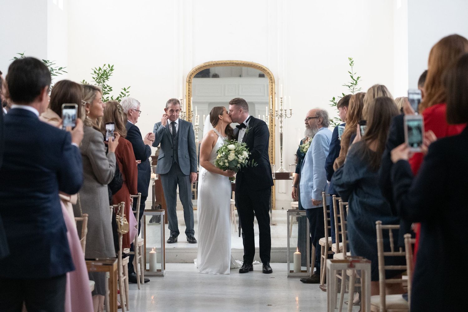 Bride and groom share first kiss at elegant white chapel wedding ceremony with guests watching from wooden chairs.