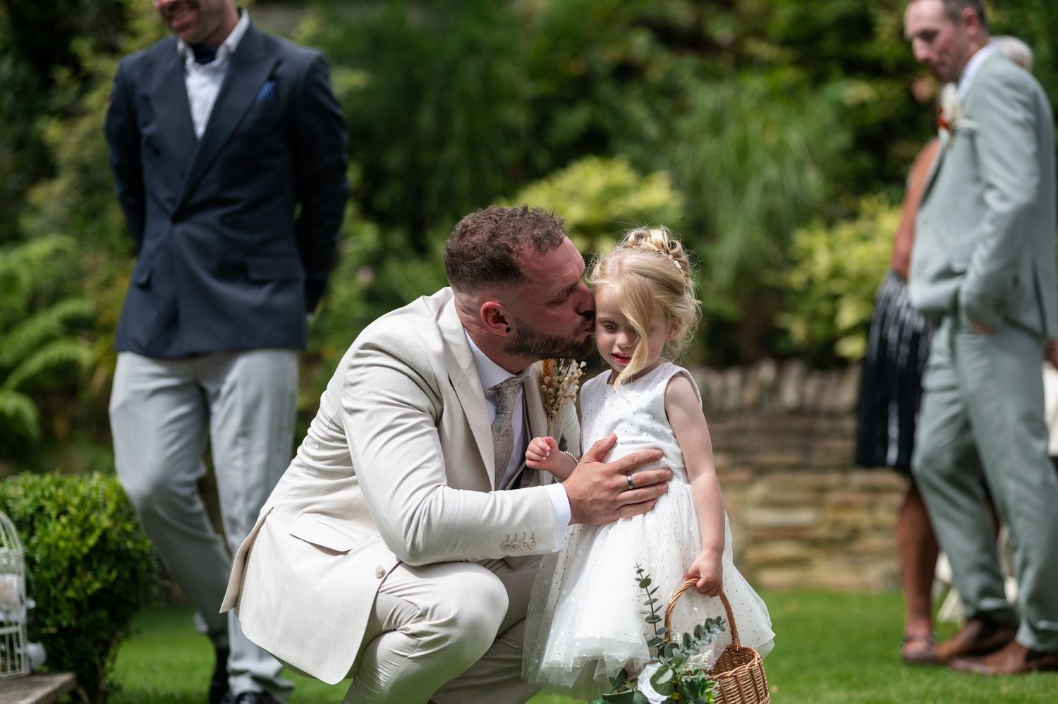 Groom in white suit crouching to kiss flower girl at outdoor wedding ceremony.