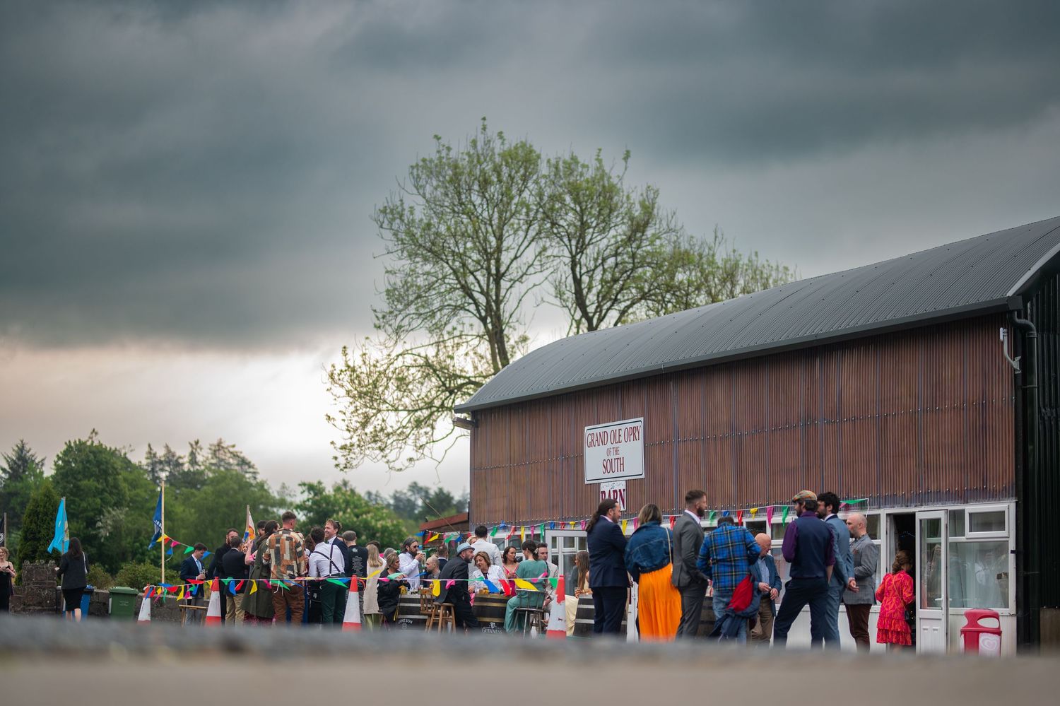 Crowd of people gathered outside a rustic brown barn venue under dramatic stormy skies at an outdoor event.