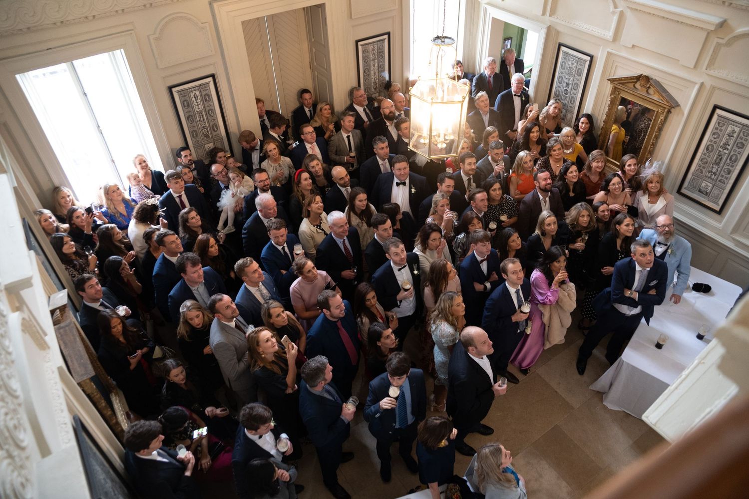 Aerial view of a large elegant gathering of guests in a grand white interior venue during a formal event.