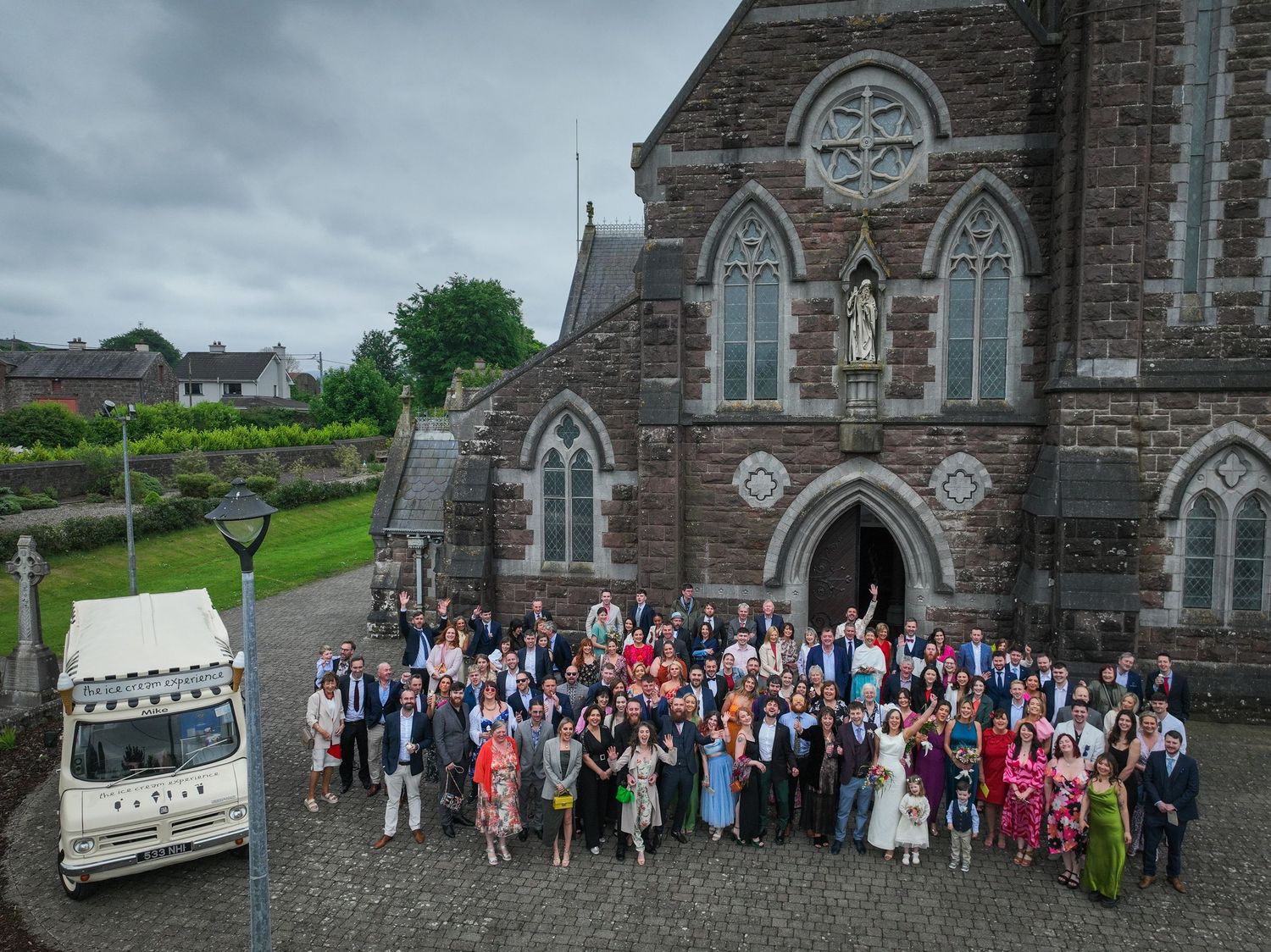 Large wedding group photo outside a historic stone church in Ireland with an ice cream van nearby.