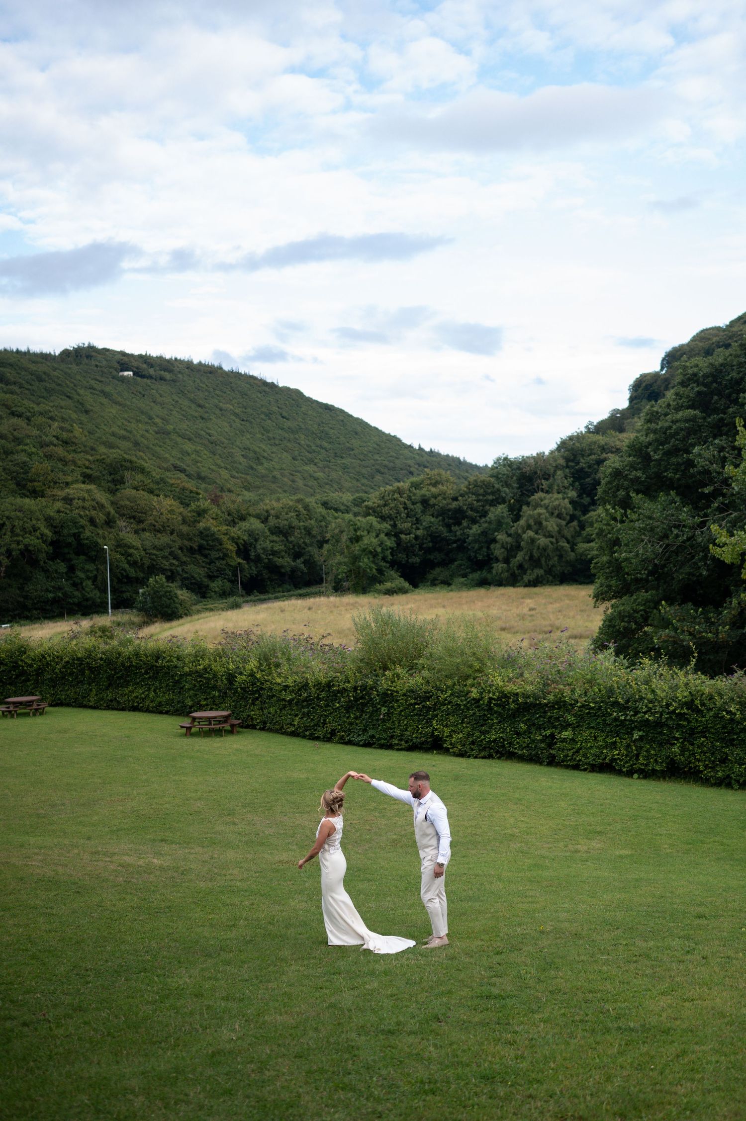 Bride and groom dancing on lush green lawn with rolling hills and trees in background on their wedding day.