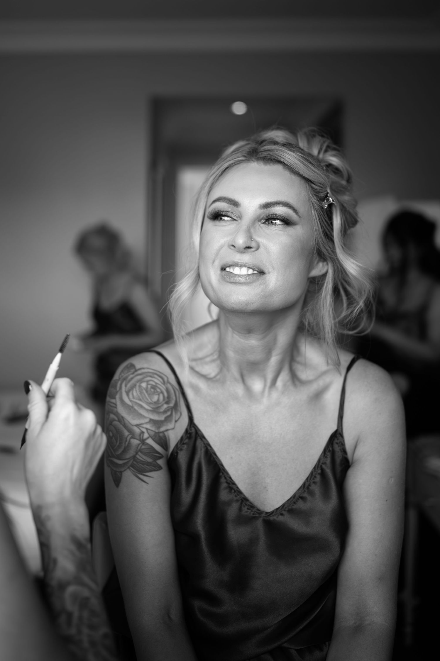 Black and white photo of a blonde woman with rose tattoo getting makeup applied at a wedding preparation.