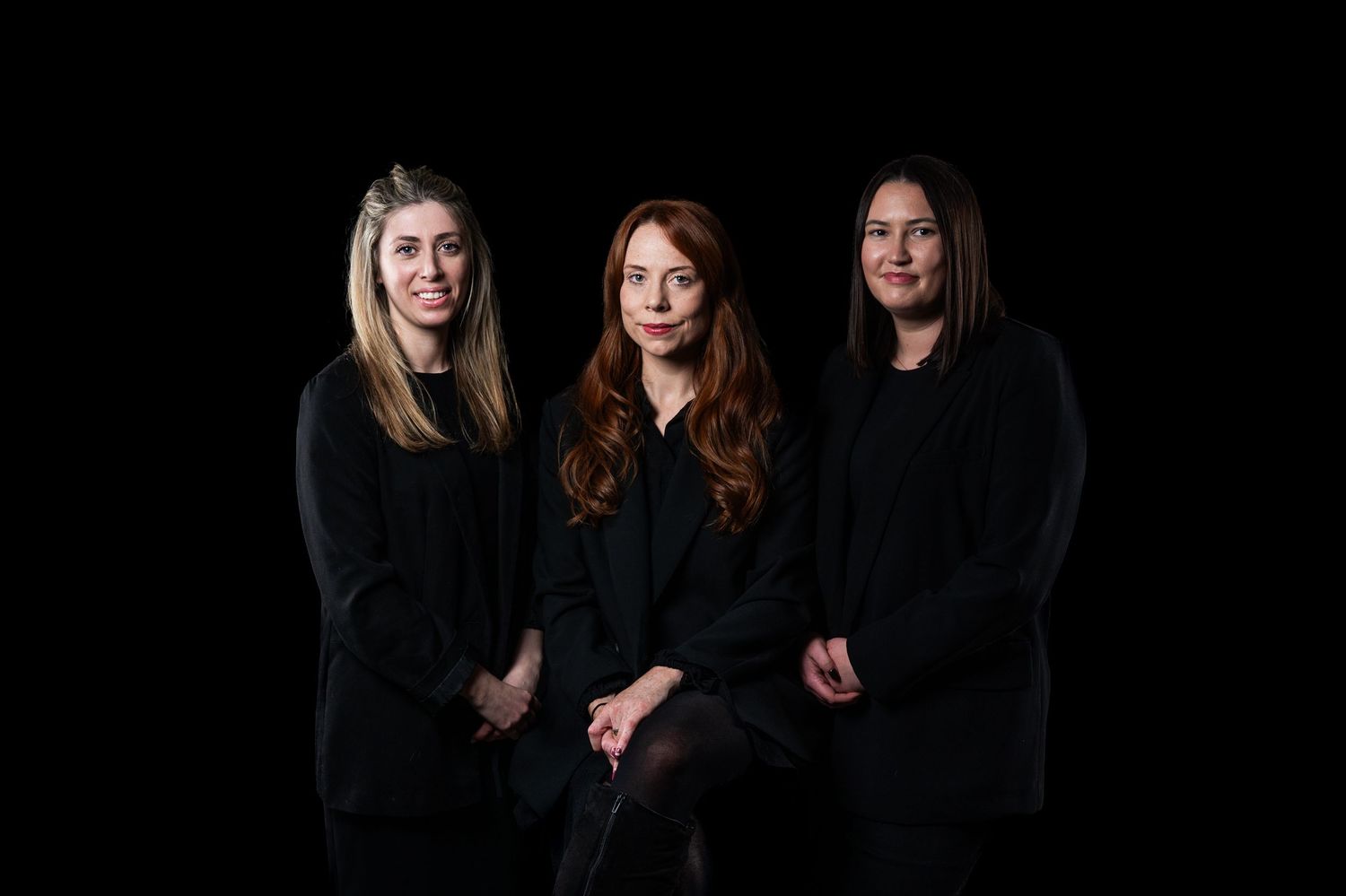 Three professional women in black attire posing together against a dark studio background.
