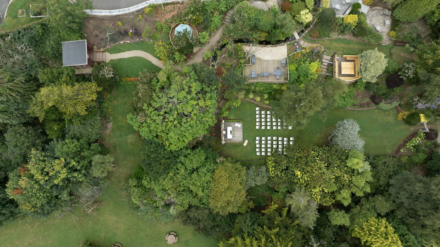 Aerial drone view of a lush garden with white chairs arranged for an outdoor ceremony.