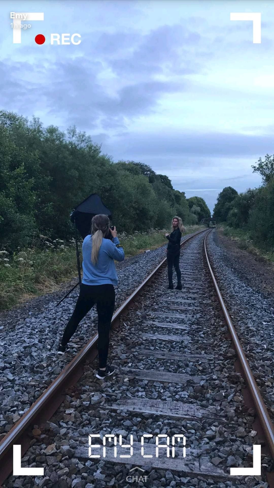 Female photographer with umbrella photographing a model standing on railway tracks on a cloudy day.