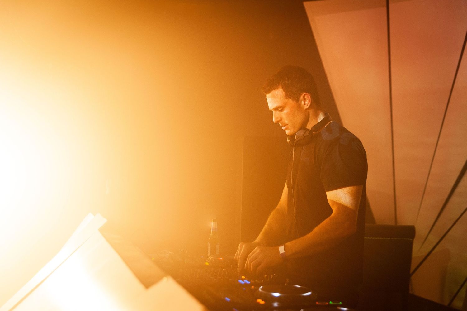Male DJ mixing at turntables bathed in warm golden stage lighting at live event.