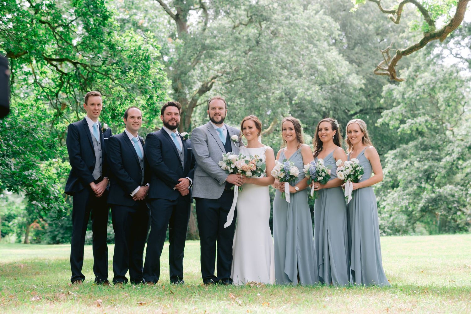 Wedding party posing outdoors with groomsmen in suits and bridesmaids in sage green dresses.