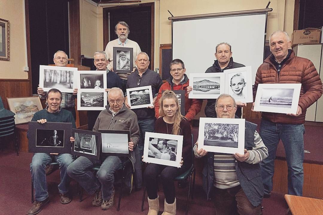 Group of photography club members proudly displaying their printed black and white photographs in a meeting room.
