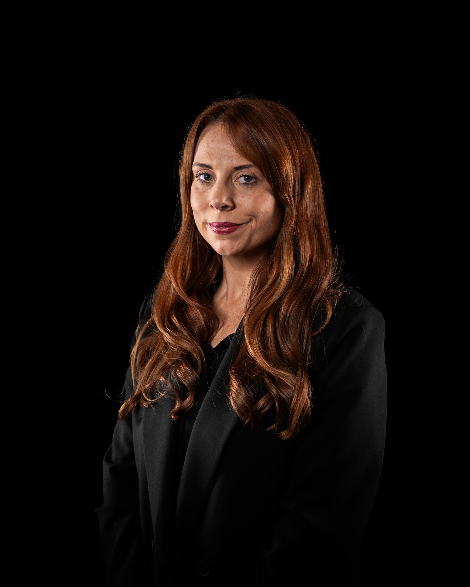 Professional headshot of a woman with long auburn hair wearing a black blazer against a dark background.