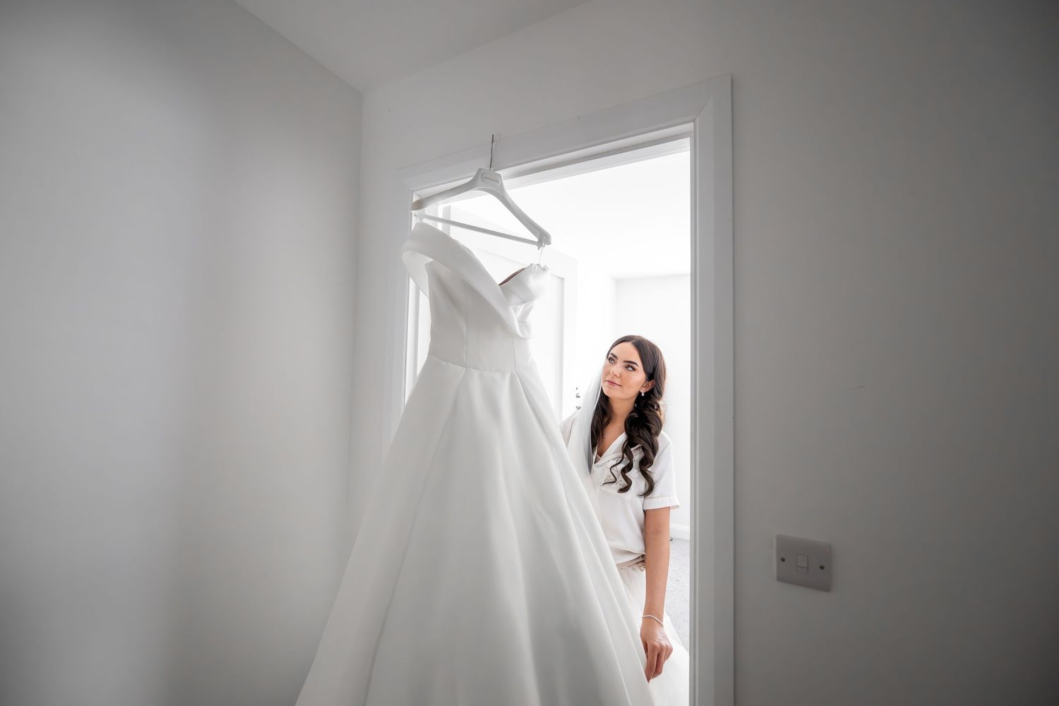 Bride in white robe admiring her elegant wedding gown hanging in a bright, airy room before the ceremony.
