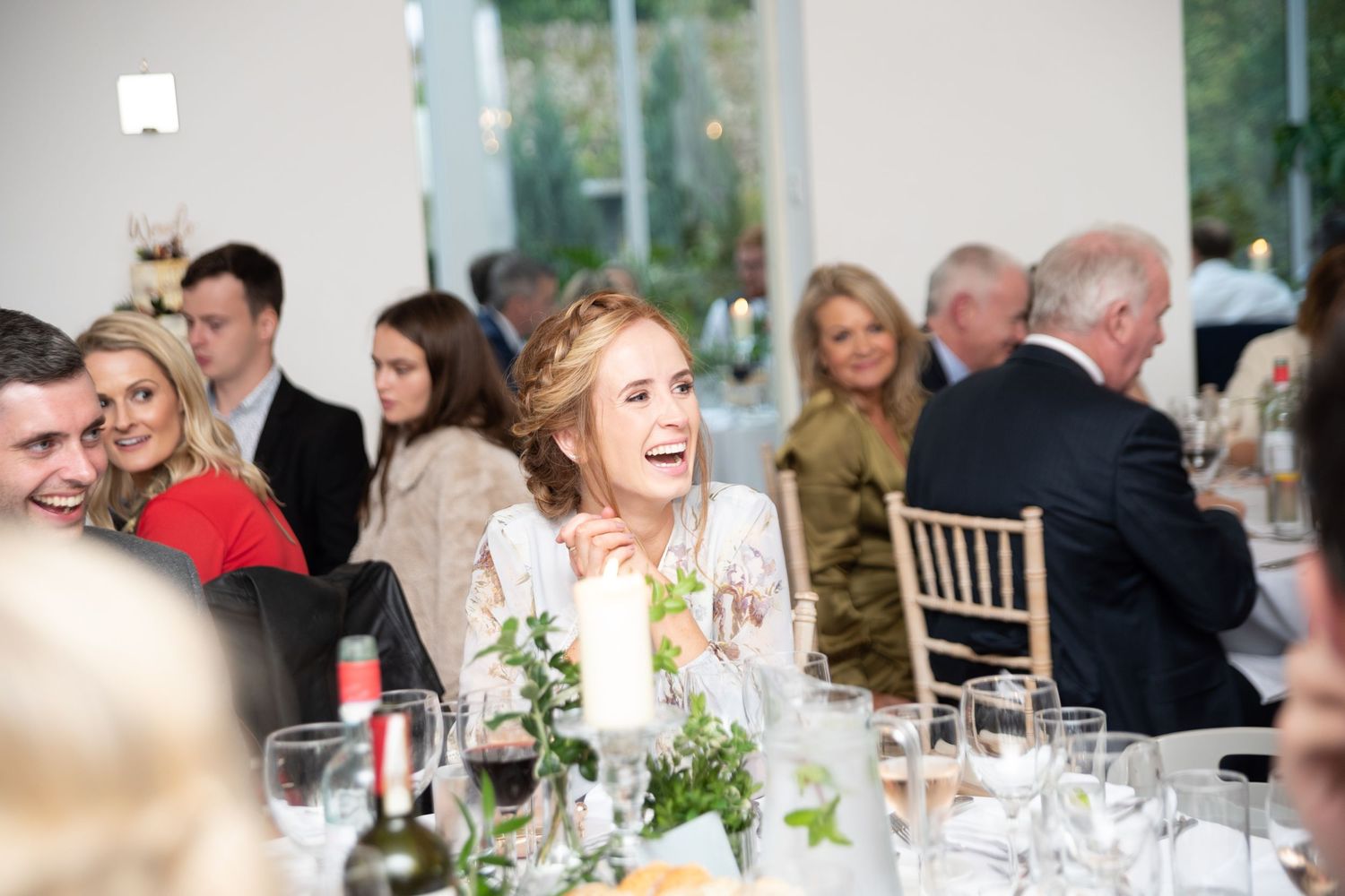Bride laughing joyfully at wedding reception dinner table with guests in elegant venue.