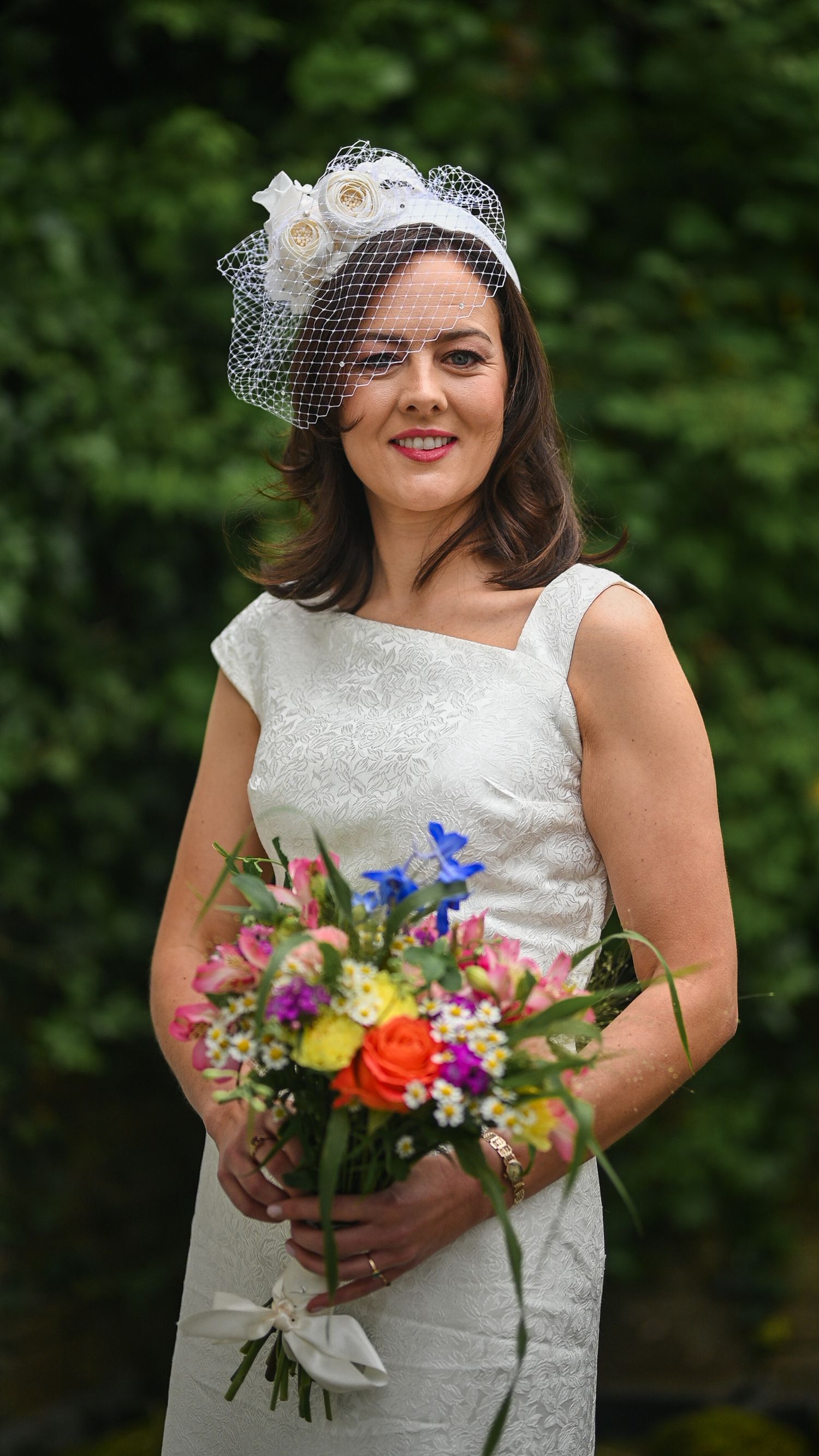Bride in white lace dress holding colorful bouquet, wearing elegant birdcage veil fascinator outdoors.
