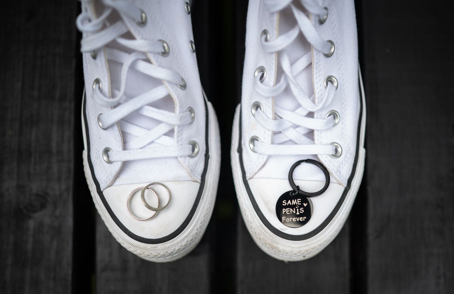 White Converse high-top sneakers with wedding rings and a personalized charm on a dark wooden surface.