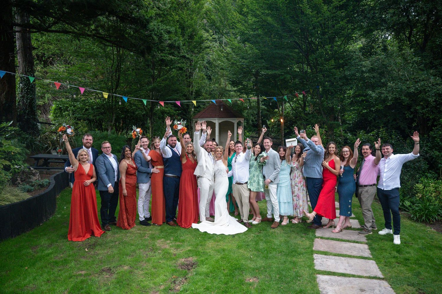 Wedding party group photo in lush green garden with colorful bunting decorations.