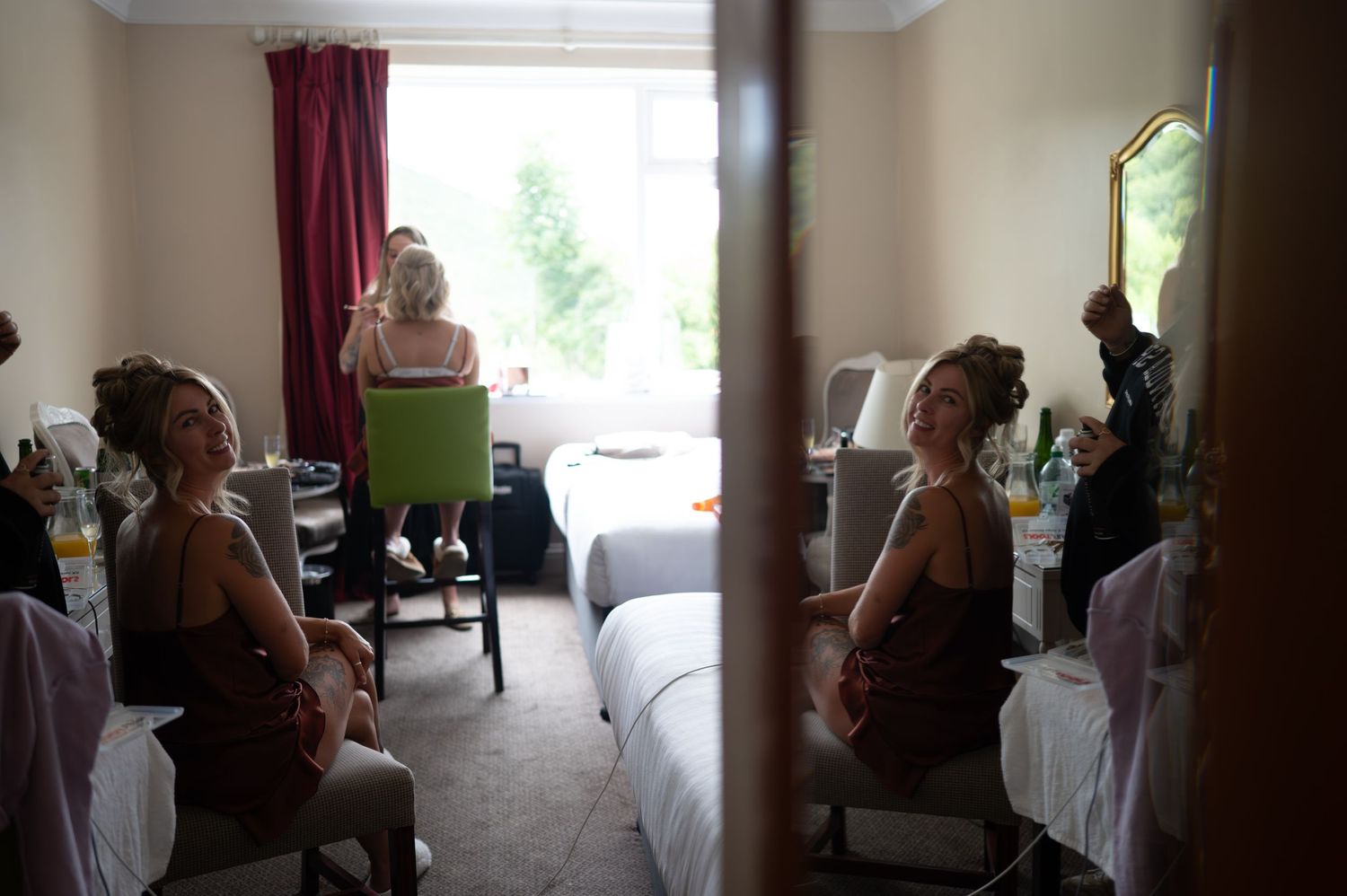 Bridesmaids in burgundy dresses getting ready in a hotel room, reflected in a mirror.