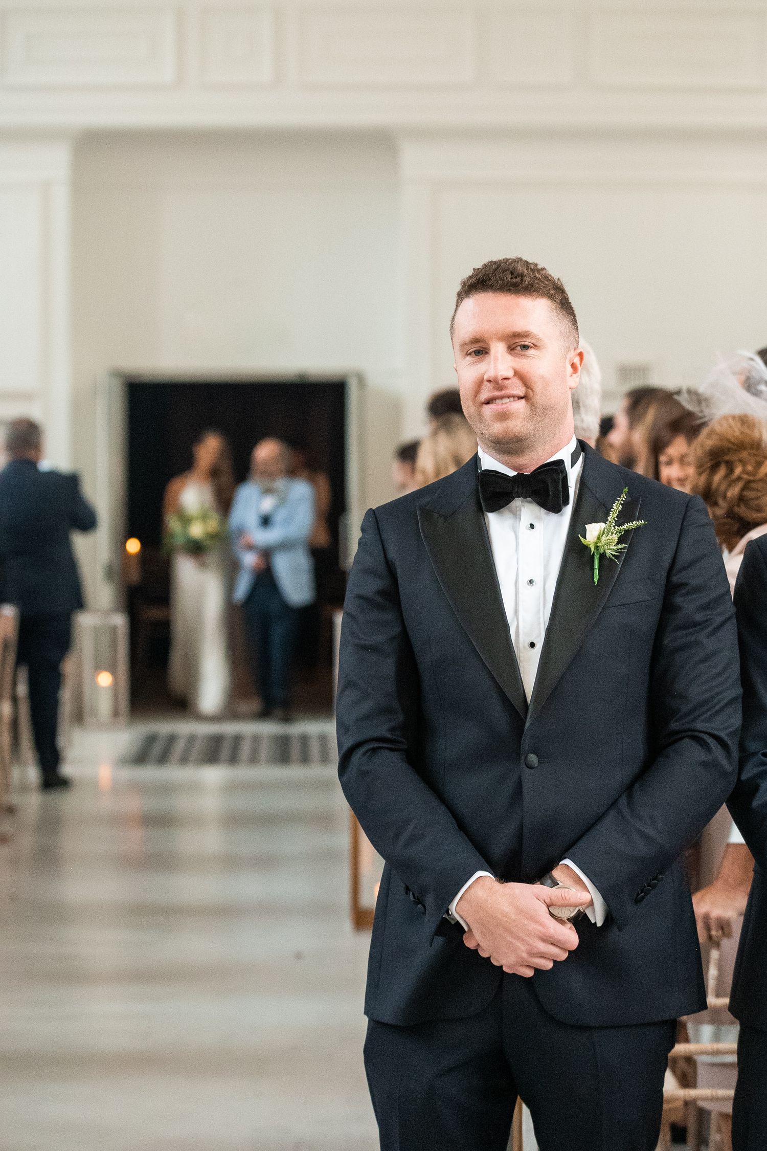Groom in black tuxedo with boutonniere smiling at altar as bride approaches down the aisle.