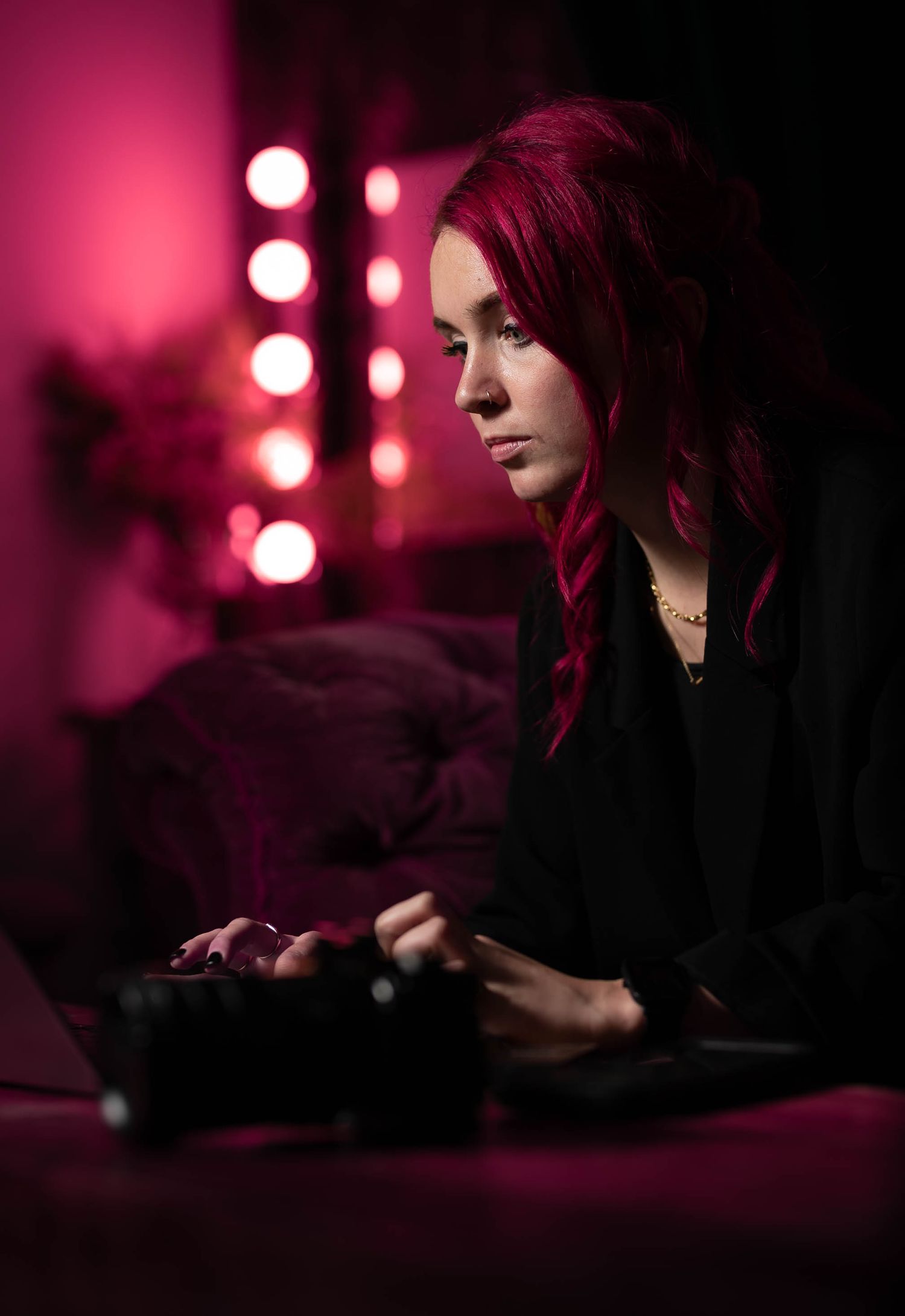 Woman with red hair focused on laptop with camera nearby in pink neon lit room.