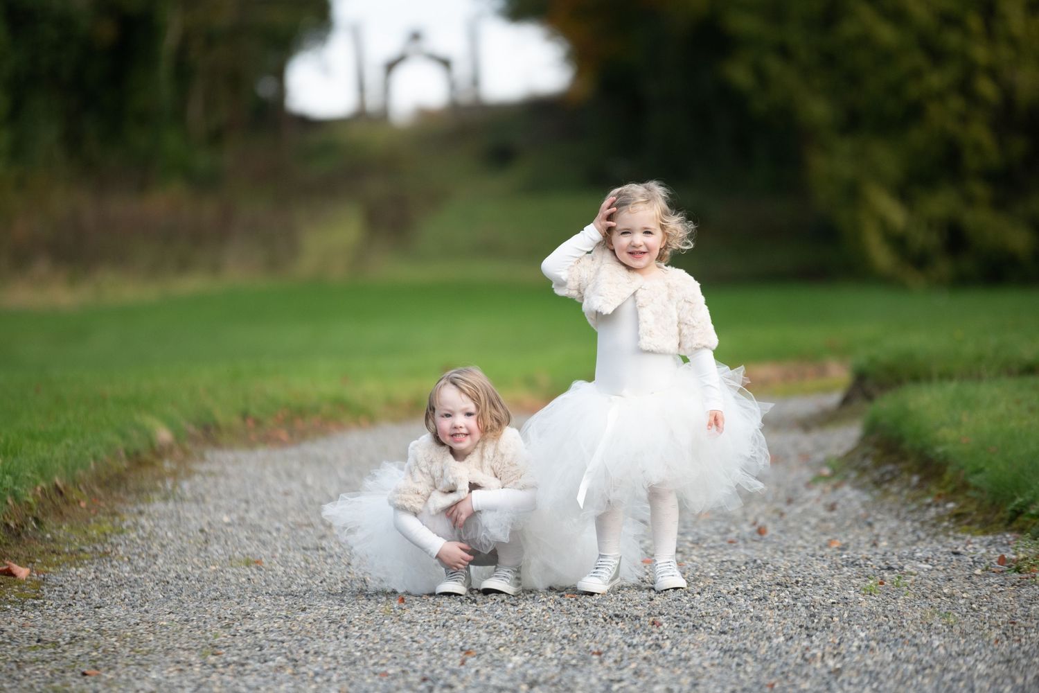 Two adorable little girls in white tulle dresses pose on a gravel path surrounded by lush green grass.