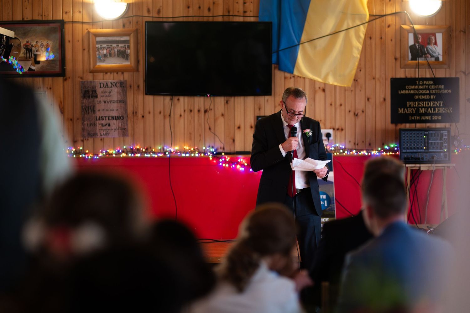 Man speaking at podium with microphone at indoor event with Ukrainian flag displayed.