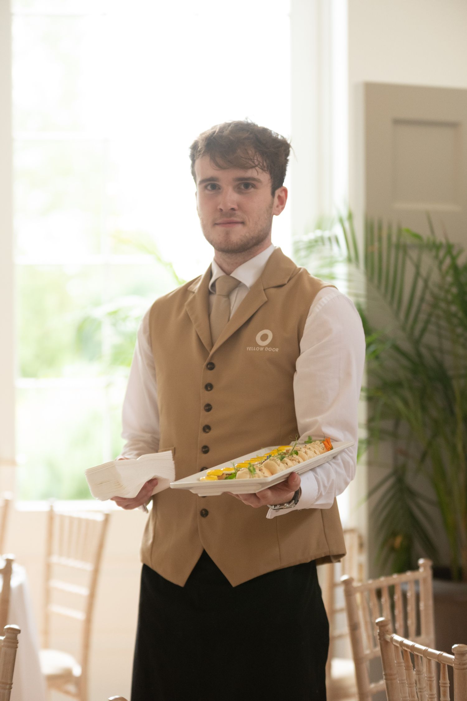 A uniformed waiter in a tan vest holds a tray of food and napkins in an elegant event venue.