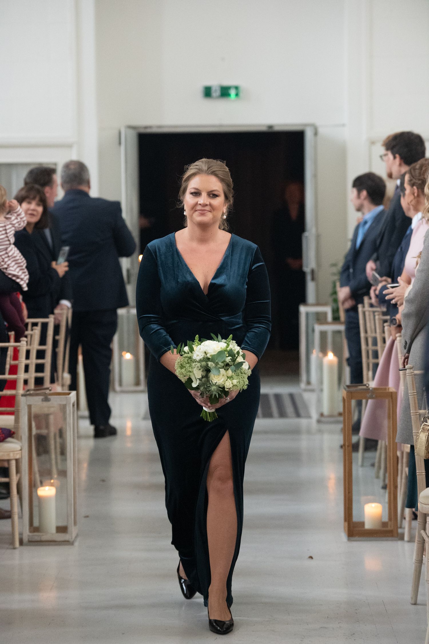 Bridesmaid in elegant dark teal velvet gown walks down aisle holding white floral bouquet at wedding ceremony.