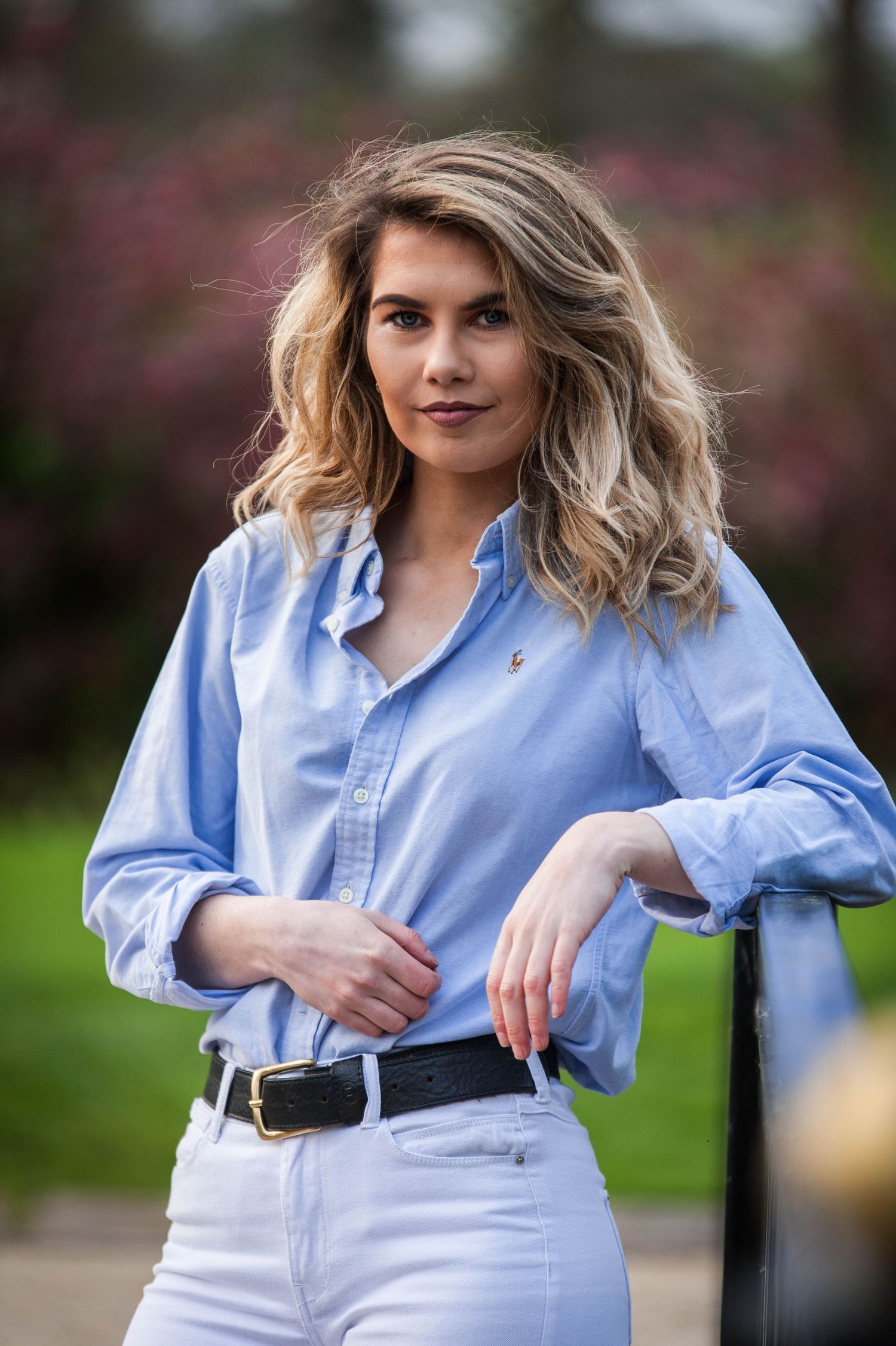 Young blonde woman in blue shirt and white jeans posing outdoors near pink flowers.