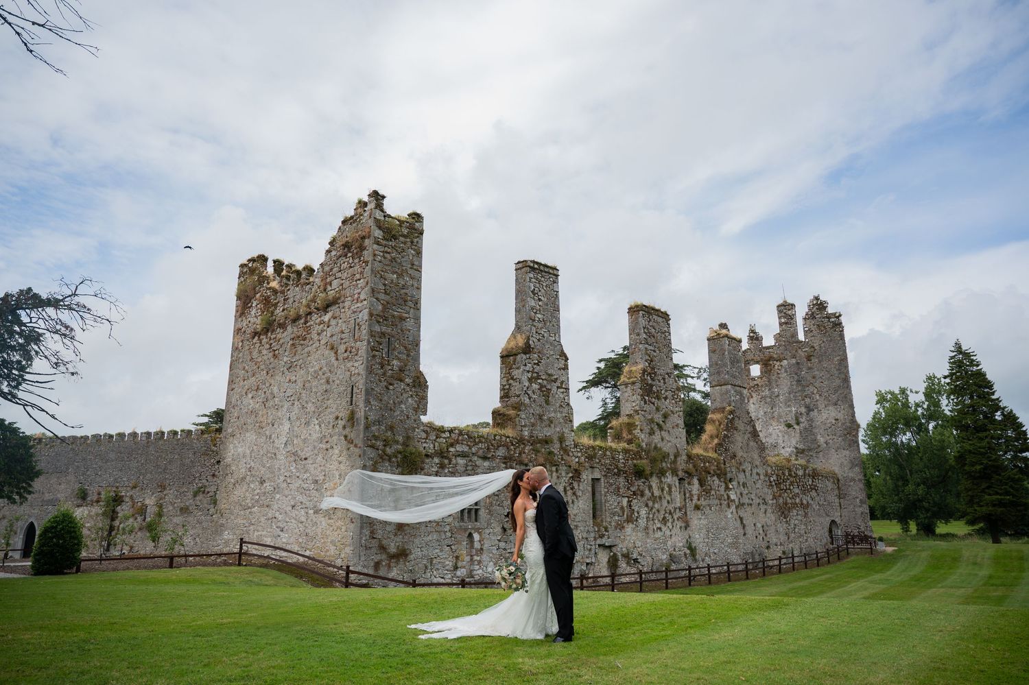 Bride and groom kissing in front of ancient castle ruins on lush green grounds on their wedding day.