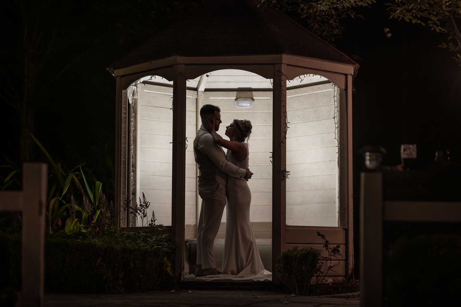 Romantic silhouette of bride and groom embracing inside illuminated gazebo at night.