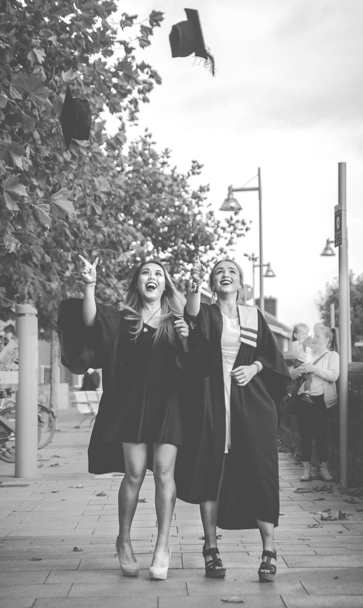 Two graduates in black robes tossing caps in air celebrating graduation outdoors in black and white.